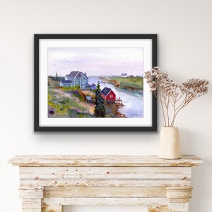 May include: Watercolor painting of a coastal landscape featuring a river, several buildings, and a red boathouse. The buildings are painted in shades of blue and gray, with green and yellow vegetation. The artwork is framed in a black frame.