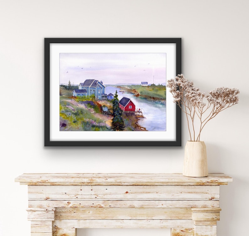 May include: Watercolor painting of a coastal landscape featuring a river, several buildings, and a red boathouse. The buildings are painted in shades of blue and gray, with green and yellow vegetation. The artwork is framed in a black frame.