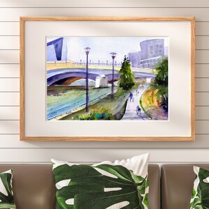 May include: A watercolor painting of a bridge over a river with people walking and biking on a path along the riverbank. The painting features a modern building in the background and trees lining the path.