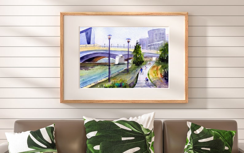May include: A watercolor painting of a bridge over a river with people walking and biking on a path along the riverbank. The painting features a modern building in the background and trees lining the path.