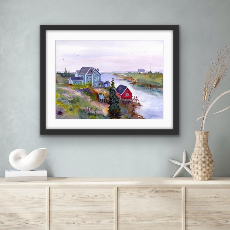 May include: Watercolor painting of a coastal scene with a red building and a blue building. The artwork is framed in a black frame and depicts a river, buildings, and a cloudy sky. The painting is displayed on a light-colored cabinet.