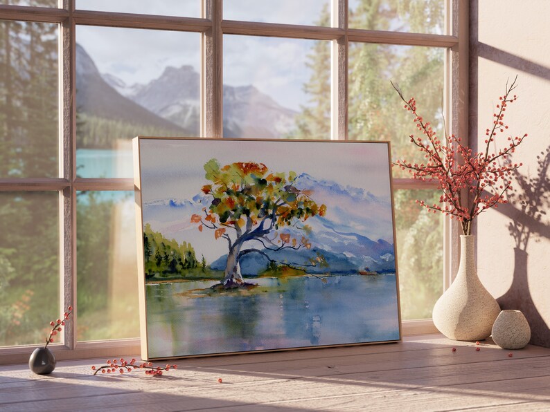 May include: A framed watercolour painting of a solitary tree with orange and yellow leaves in a mountainous landscape. The tree is in the foreground, with the mountains in the background. The water is a pale blue colour, and the sky is a light grey colour.