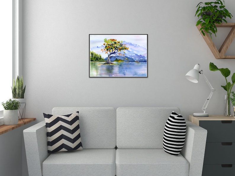 May include: Watercolour painting of a solitary tree with orange and green leaves on a small island in a lake. The lake is surrounded by mountains and a blue sky.