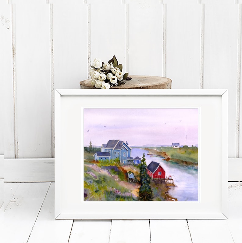 May include: A framed watercolor painting depicts a coastal scene with blue houses, a red building, and a river. The artwork is set in a white frame, with a small bouquet of white flowers on a wooden slice above it.