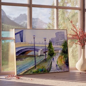 May include: A watercolor painting of a bridge over a river with mountains in the background. The painting is framed in a wooden frame and is hanging on a wall.