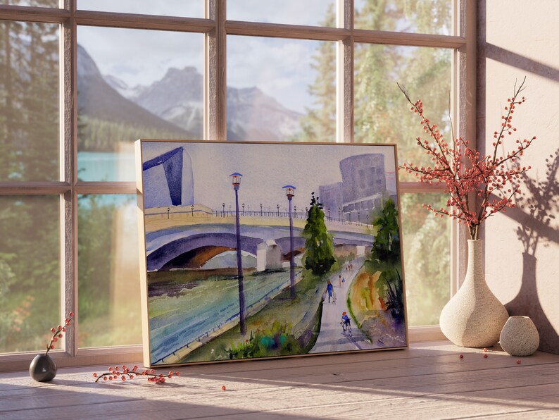 May include: A watercolor painting of a bridge over a river with mountains in the background. The painting is framed in a wooden frame and is hanging on a wall.