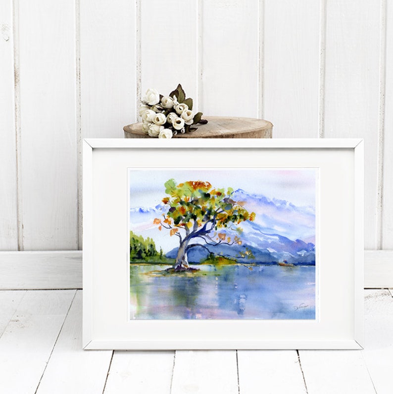 May include: A framed watercolour painting of a solitary tree with orange and yellow leaves standing in a lake with mountains in the background.