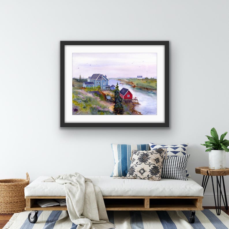 May include: A framed watercolor painting depicts a coastal scene with a river, houses, and a tree. The houses are painted in shades of blue and red. The artwork is displayed on a white wall above a white couch with decorative pillows.