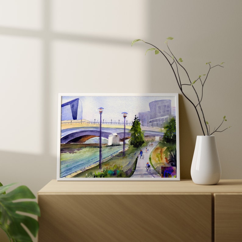 May include: Watercolor painting of a bridge over a river with people walking on a path along the riverbank. There are streetlights on the bridge and trees along the riverbank. The painting is framed in white.