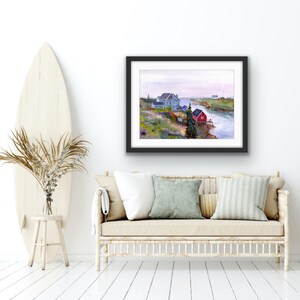 May include: A framed watercolor painting depicts a coastal scene with a red building, several blue buildings, and a body of water. The artwork is displayed above a beige sofa with pillows, next to a surfboard and a vase of dried plants.