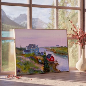 May include: A framed watercolor painting of a coastal village scene. The artwork depicts houses in shades of blue and red along a river, with a backdrop of mountains and a soft, pastel sky. The painting is displayed on a wooden surface near a window.