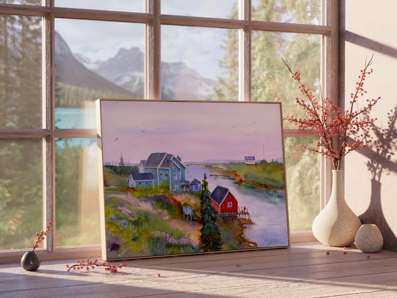 May include: A framed watercolor painting of a coastal village scene. The artwork depicts houses in shades of blue and red along a river, with a backdrop of mountains and a soft, pastel sky. The painting is displayed on a wooden surface near a window.