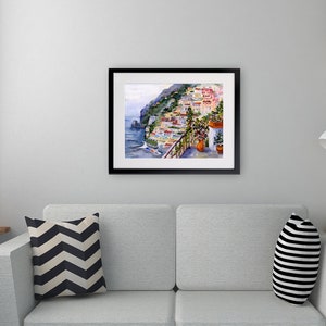 May include: A watercolor painting of a colorful Italian village on a cliffside overlooking the sea. The painting is framed in black and is hanging on a white wall above a gray couch.