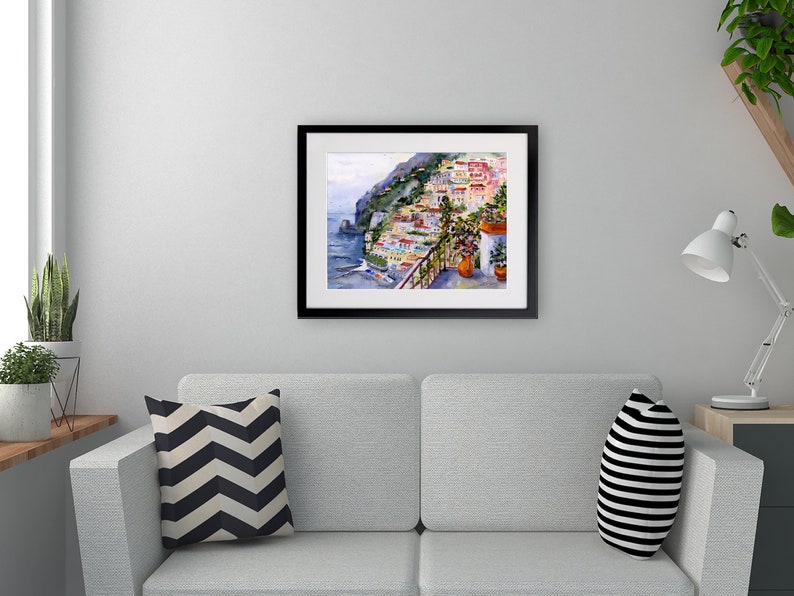 May include: A watercolor painting of a colorful Italian village on a cliffside overlooking the sea. The painting is framed in black and is hanging on a white wall above a gray couch.