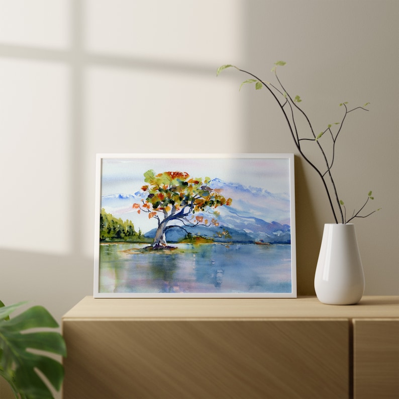 May include: Watercolour painting of a solitary tree with orange and yellow leaves standing in a lake with mountains in the background.