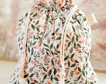 Puede incluir: Una bolsa de cordón blanco con un patrón floral con flores rosas y hojas verdes. La bolsa tiene dos botones de madera en los cordones.