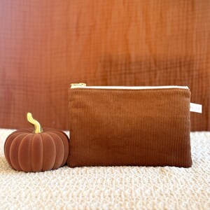 Pode incluir: Uma bolsa de veludo cotelê marrom com zíper dourado e uma pequena abóbora decorativa com haste dourada. A bolsa e a abóbora estão em uma superfície creme com um fundo marrom.