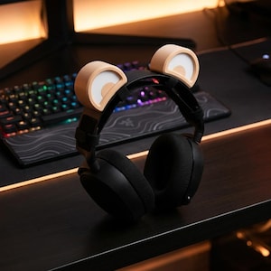 Könnte beinhalten: Schwarzer Gaming-Kopfhörer mit hellbraunen Bärenohranbauten. Der Kopfhörer liegt auf einem schwarzen Schreibtisch, im Hintergrund befinden sich eine Tastatur und ein Monitor. Die Ohranbauten haben ein weißes und hellbraunes Design.