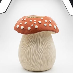 Puede incluir: Un jarrón de cerámica con forma de hongo. El sombrero es de color marrón rojizo con puntos blancos, y el tallo es de color crema con líneas verticales texturizadas. Un artículo decorativo.