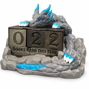 Könnte beinhalten: Eine graue Drachenfigur mit leuchtend blauen Akzenten an Hörnern, Augen und Krallen auf einem felsigen Sockel. Sie enthält einen rechteckigen Block mit der Zahl 022 und dem eingravierten Text BOOKS READ THIS YEAR in dunkler Metalloptik.