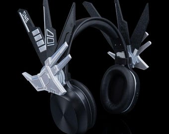 May include: Black headphones with a futuristic design. The headphones have a padded headband and earcups. The earcups are circular and have a silver trim. The design includes angular, geometric accents in black and silver, with the word "ONE" visible.