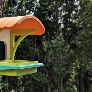 May include: A brightly coloured bird feeder attached to a tree. It features an orange roof, white walls, and green and yellow supports. A black camera is visible inside. The feeder has a tray for food and is set against a backdrop of green trees.