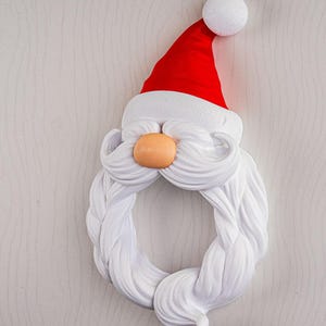 May include: A whimsical Santa Claus wreath with a red hat, white beard, and orange nose. The wreath is designed in a circular shape, perfect for holiday decor. The hat features a white pom-pom at the top. The beard is a braided design.