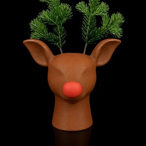 Puede incluir: Una cabeza de reno de cerámica marrón con una nariz roja brillante y ramitas verdes como cornamenta. El reno tiene orejas grandes y está sobre un fondo negro, evocando un tema festivo navideño.