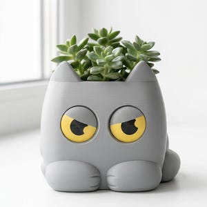 Puede incluir: Una maceta gris con forma de gato con ojos amarillos y negros, que contiene una planta suculenta verde. La maceta tiene un diseño caprichoso, con rasgos redondeados y orejas pequeñas. La planta es de un verde vibrante, que contrasta con el gris de la maceta.