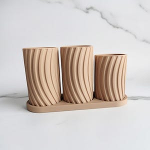 Puede incluir: Un conjunto de tres macetas de cerámica beige con una bandeja a juego. Cada maceta cilíndrica presenta un diseño vertical acanalado. Las macetas están dispuestas en fila, variando en altura, y son adecuadas para plantas pequeñas o suculentas.