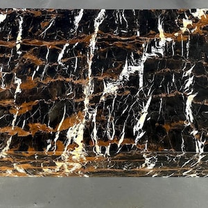 May include: Rectangular black marble table with gold and white veining. The table has a polished surface and a solid, block-like construction. The marble pattern creates a striking contrast of colors and textures, suitable for home decor.
