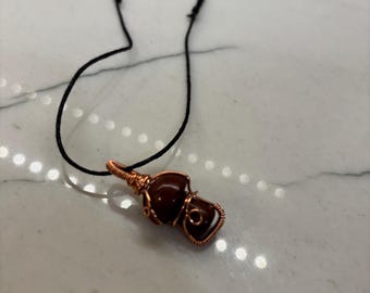 以下が含まれることがあります： 黒い紐に、銅線で巻かれた茶色の宝石のペンダントがぶら下がっている。