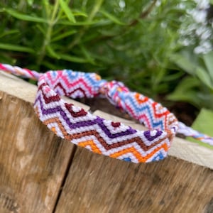 May include: A colorful friendship bracelet with a chevron pattern in shades of white, orange, brown, purple, pink, and blue.