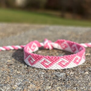 May include: A pink and white friendship bracelet with a geometric pattern. The bracelet is made of woven threads and is lying on a textured gray surface. A matching pink and white cord is visible in the background.