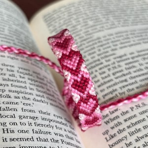 May include: A handmade friendship bracelet in shades of pink and white, featuring a chevron pattern. The bracelet is resting on the open pages of a book, showcasing its intricate design and craftsmanship. A perfect gift.