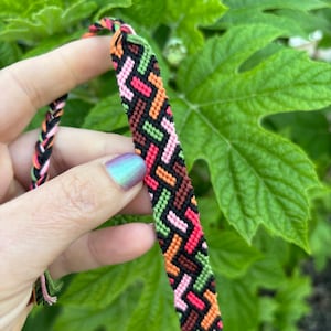 May include: A colorful friendship bracelet with a geometric pattern in shades of pink, green, orange, black, and brown.