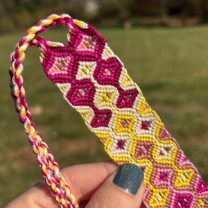 May include: A pink and yellow friendship bracelet with a diamond pattern.