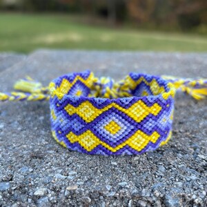 May include: Handmade friendship bracelet in shades of purple and yellow. The woven bracelet features a geometric diamond pattern. The ends of the bracelet have braided cords with yellow tassels. This accessory is perfect for adding a pop of color.