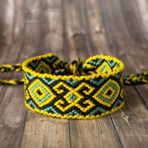 May include: A woven bracelet with a geometric pattern in yellow, black, and teal. The bracelet is displayed on a wooden surface. The design features repeating diamond and square shapes.