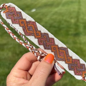 May include: Handmade friendship bracelet with a geometric pattern in shades of orange, purple, and brown on a white background. The bracelet has a braided cord and a small tassel. The bracelet is held against a green background.