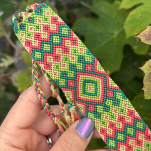 May include: A colourful friendship bracelet with a geometric pattern in green, coral, and beige. The bracelet is held up, showing the intricate woven design and braided ties.