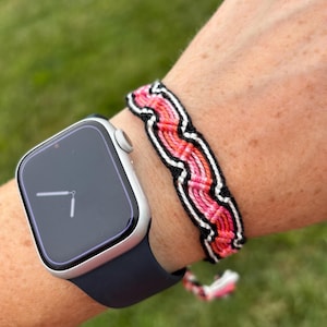 May include: A silver smartwatch with a dark blue band is next to a woven friendship bracelet. The bracelet features a pattern of pink, red, black, and white threads. The bracelet is on a wrist.