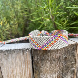 May include: A woven friendship bracelet with a geometric pattern in shades of pink, orange, yellow, and blue on a beige background.
