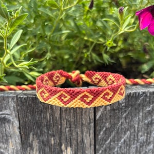 May include: A handmade friendship bracelet with a geometric pattern in red and yellow. The bracelet is made of woven threads and is displayed on a wooden surface with a green leafy background.