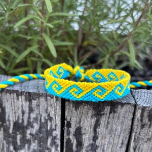 May include: A handmade friendship bracelet in yellow and turquoise blue, featuring a geometric pattern. The bracelet is displayed on a weathered wooden surface, with green foliage in the background. The bracelet measures approximately 0.5 inches wide.