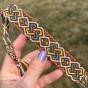 May include: A handmade friendship bracelet featuring a geometric pattern in brown, yellow, teal, and white. The bracelet is held in a hand and has a braided tassel. The design is intricate and colorful.