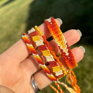 May include: A collection of handmade friendship bracelets in shades of orange, yellow, and cream. The bracelets feature various woven patterns and textures, with some having a braided design. The bracelets are displayed against a blurred green background.