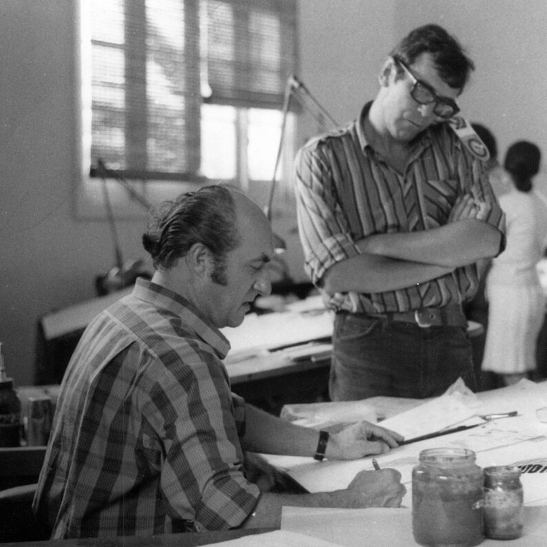 May include: Two people are working at a drafting table. One person is wearing a striped shirt and has their arms crossed. The other person is wearing a plaid shirt and is drawing on a piece of paper. There are jars of paint on the table.