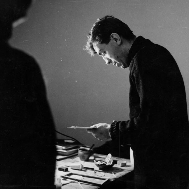 May include: A black and white photo of a person looking at a small object on a table. The person is wearing a dark sweater and has short, dark hair. The table is covered with a white cloth and has several small objects on it.