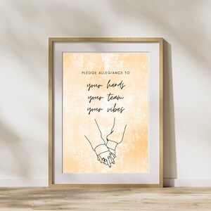 May include: Framed art print with a light orange watercolour background. The print features the text "Pledge Allegiance To" above the words "your hands your team your vibes" in a handwritten style. Below the text is a line drawing of two hands holding each other.